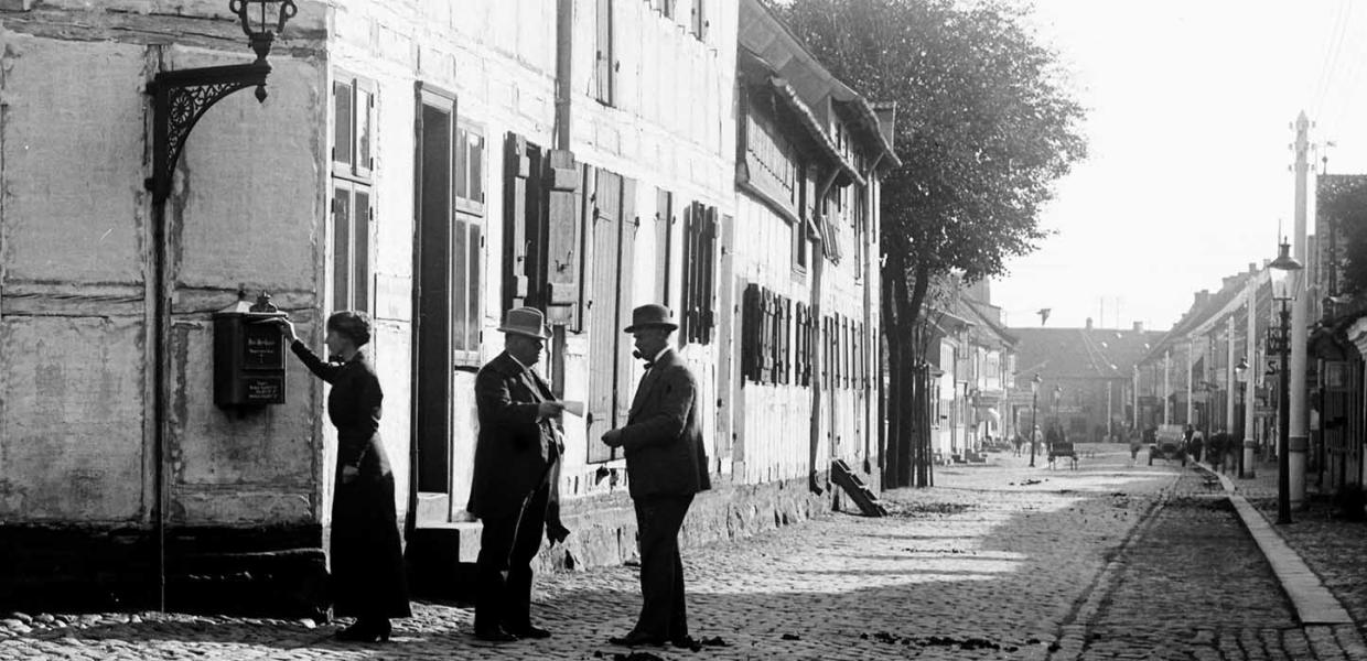En kvinde står ved siden, og to mænd med bowlerhatte på taler sammen ved Trægården i gamle dage