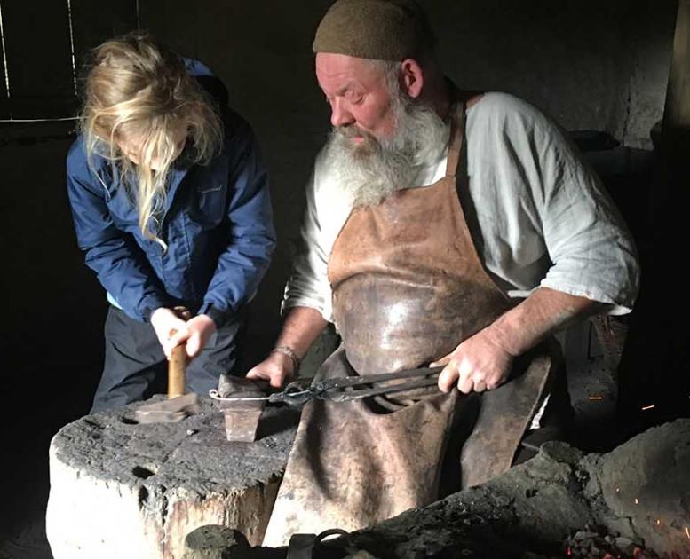 Jernaldersmeden holder metallet, som pigen skal hamre på